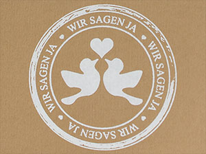 Weißer Stempelabdruck auf braunem Kraftkarton, den Schriftzug wir sagen ja, sowie zwei turtelnde Tauben mit weißem Herz darstellend.